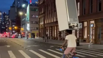 A man balances a refrigerator on his head while cycling down a busy street