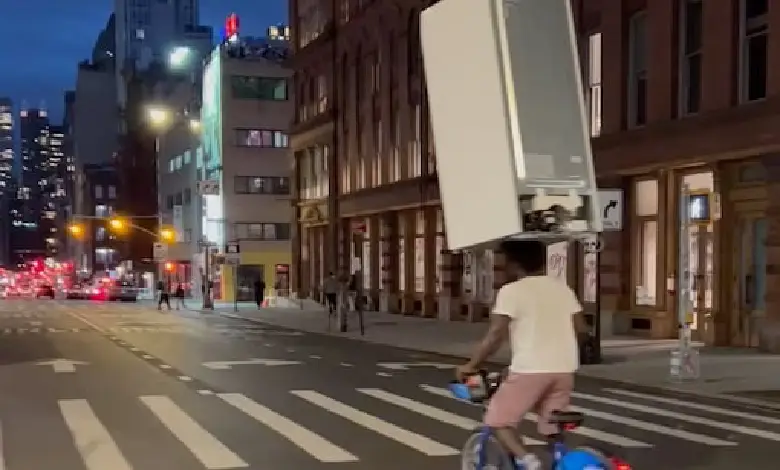 A man balances a refrigerator on his head while cycling down a busy street