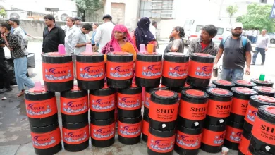 People gathered under a temporary shade arrangement, receiving water and snacks from volunteers, near a hospital building.