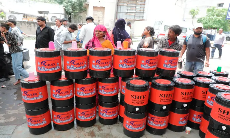 People gathered under a temporary shade arrangement, receiving water and snacks from volunteers, near a hospital building.